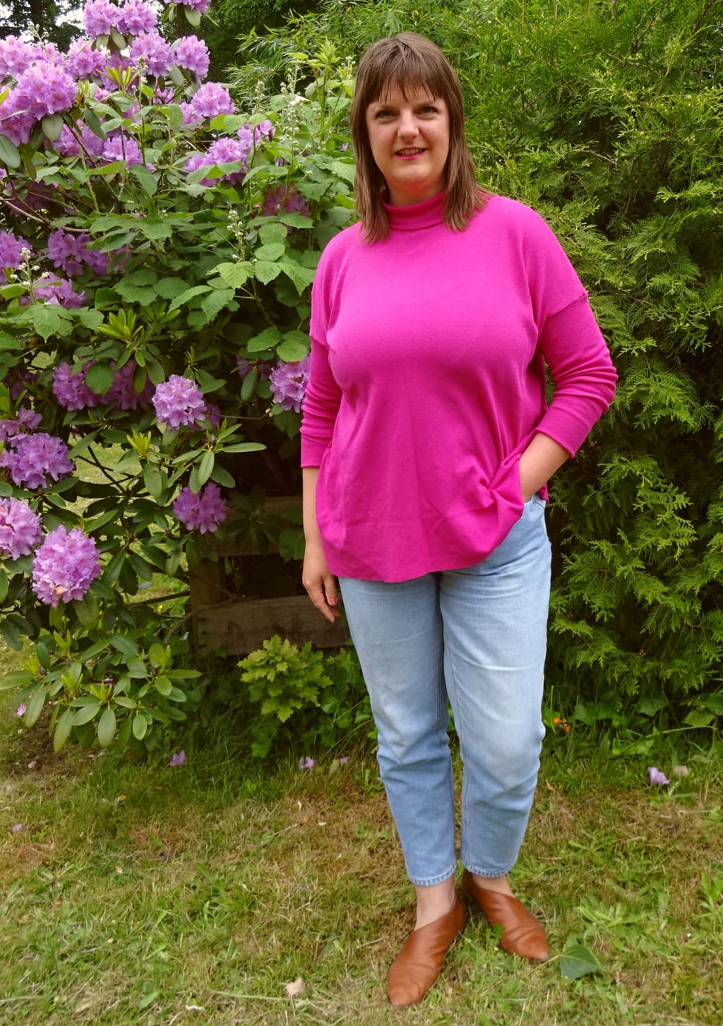 Tine Sews posiert in einem pinken Monroe Turtleneck vor einem pinken Rhododendron