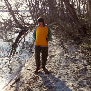 Frau geht einen Strand entlang und trägt ein buntes Sweatshirt mit Taschen