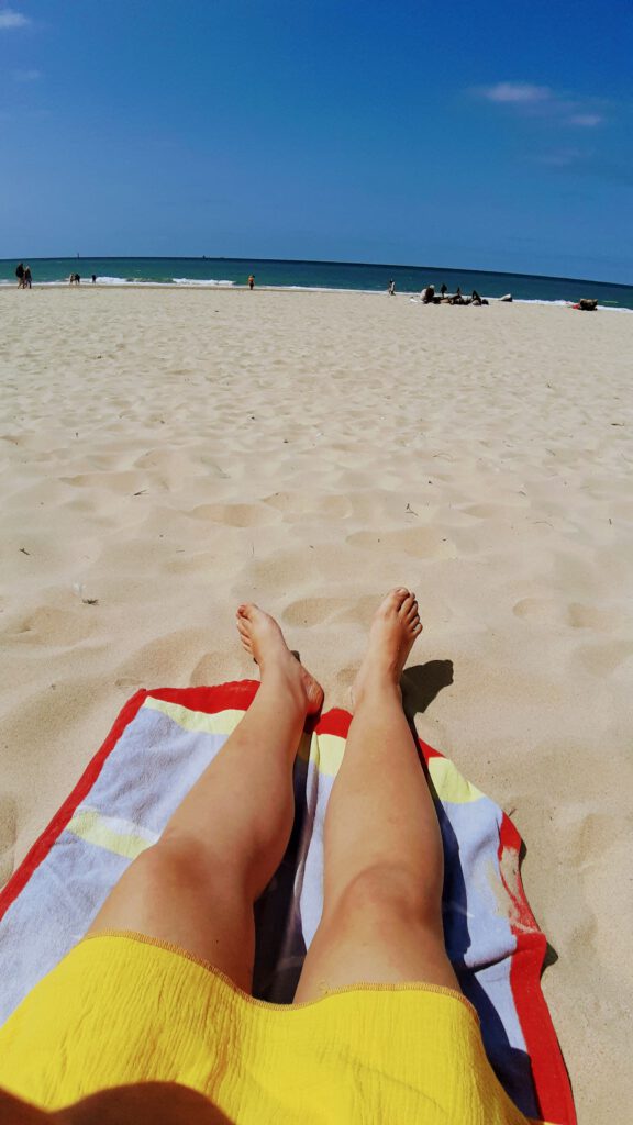Tine liegt am Strand von Westerland und man sieht den gelben Surfponcho und ihre Beine. Im Hintergrund sieht man die Nordsee.